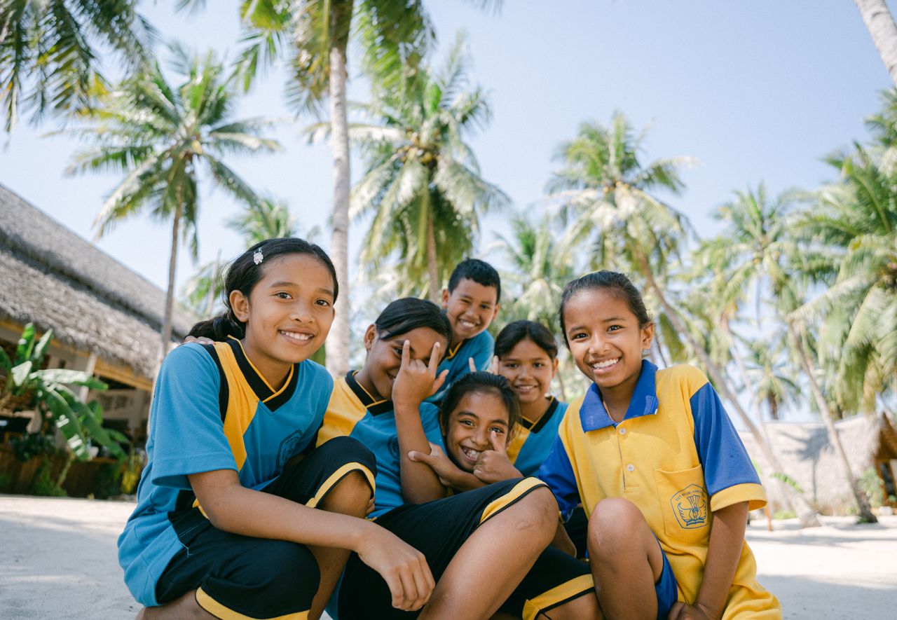 Group of smiling children in uniforms playfully posing under palm trees on a sunny day.